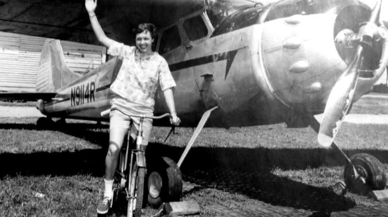 Wally Funk waving in front of an aircraft while riding a bicycle